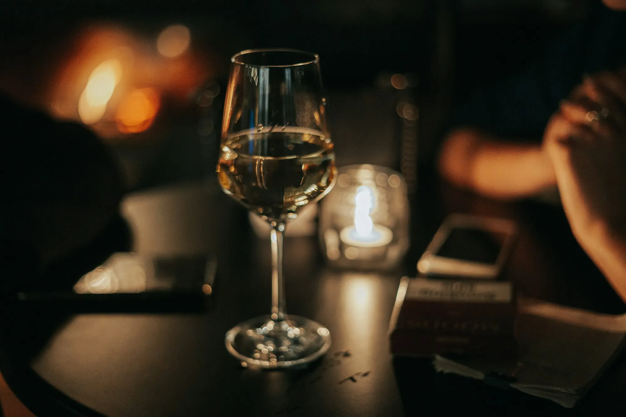 Taça de vinho em primeiro plano num restaurante à meia-luz, com alguém desfocado ao fundo.
